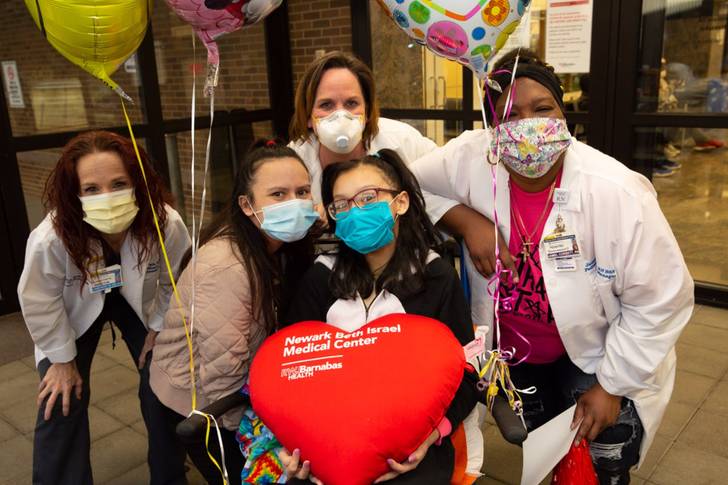 Jissel Rosario sits in the center of medical workers who helped her; all are wearing masks, and she's holding balloons and a big heart pillow from the hospital